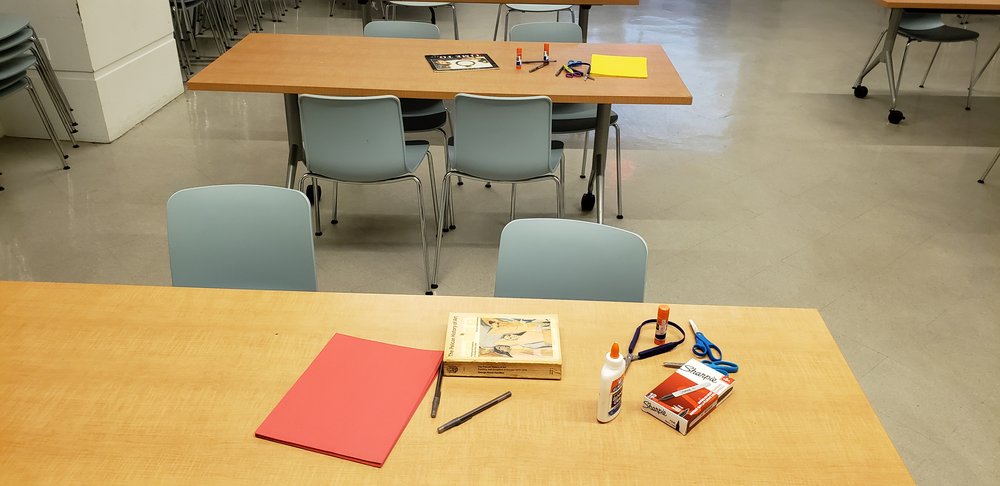 Two tables with books and supplies on them. The front table has both adaptive and regular scissors, glue, glue sticks pens, sharpies, red paper, and thick book. The back table has a children’s book, yellow paper, glue, glue sticks, and scissors. Both tables have gray chairs around them.