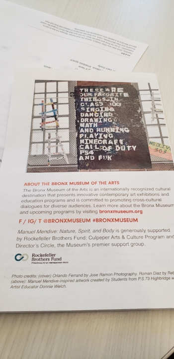 Small paper flyer on a white table. The flyer has a photograph of the large, mural, praise poem in situ at the school against a brick wall with other art projects surrounding it. Below the photo is a description of the Bronx Museum, it’s social media content, and the funders/donors.