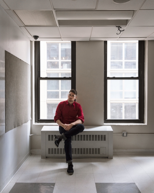 (Photo by  Julianne Nash ) Donnie Welch, young caucasian male, sits on radiator by the window of a empty classroom. He’s in a red shirt, jeans, and sneakers, sitting with one leg crossed. The gray and white tile floor and white board of the classroom are visible in the foreground.