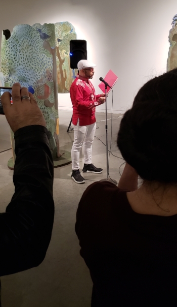 Afro-Cuban Poet Orlando Ferrand reads into a microphone from a red folder. He is wearing all white beret, glasses, a red shirt, white pants and black sneakers. In the foreground are audience members holding phones up to film and take pictures. The background is the Mendvie exhibit. No single piece of art is all in frame, but the nature elements of Mendive are clear with trees, leaves, roots, birds, jungle like plants, and human figures all visible in blues, greens, red, yellows, and browns.