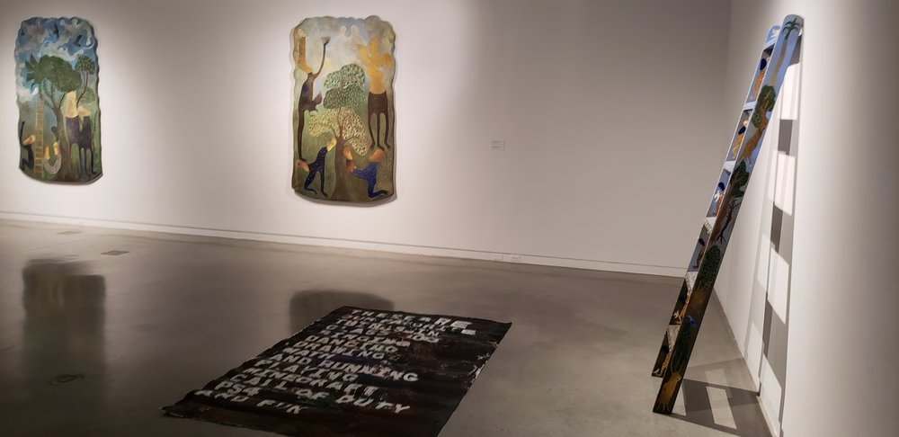 Large, mural poem is load out on a concrete gray exhibit floor. The mural poem itself is colorful, with white letters popping out through greens, purples, reds, blues, and yellows, to form words. The angle of the photo makes it difficult to see the start of the poem, though the lower words “ And running / playing/ minecraft / call of duty/ PS4/ and fun” are visible. In the background is the Manuel Mendive exhibit full of natural imagery with trees, leaves, roots, birds, jungle like plants, and human figures all visible in blues, greens, red, yellows, and browns. To the right is one of Mendive’s ladders also featuring the nature themes in smaller detail along the side and on the steps.