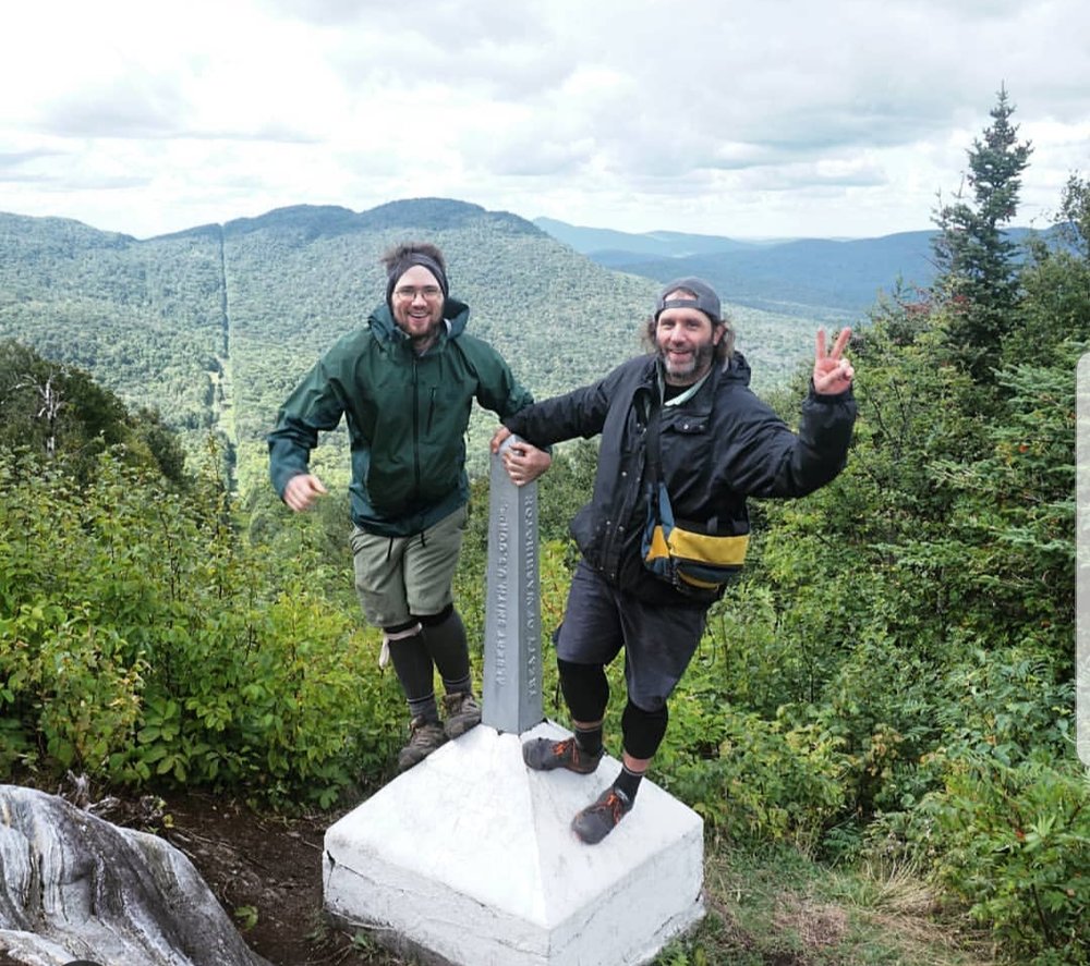 Donnie Welch and Jonathan Welch, two bearded young men, stand on the Canadian-US border in Vermont. The two are standing on a silver, spiked monument with a white base which denotes the border line. Behind them can be seen mountains and forests with a clear cut line for the border. The looks on their faces are ones of excitement and joy