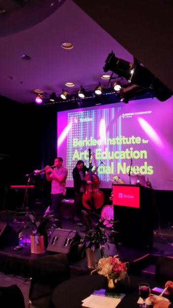In purple-hued stage lights two musicians play. A younger black male in a short sleeve gray shirt and jeans plays trumpet with a younger white male with long hairs in a black cardigan and dark jeans plays stand-up bass. They’re both on stage with large monitors, a drum set, and key board around them a and screen bearing the conference title behind them