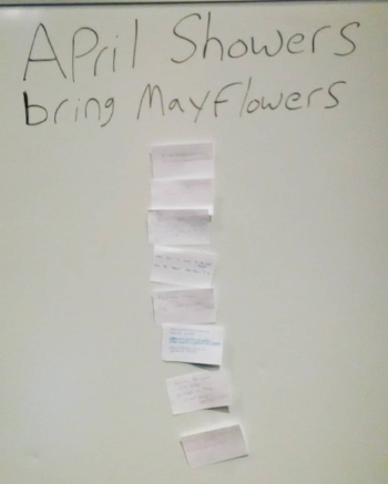 “April Showers / bring Mayflowers” is written in black lettering on a white board. Below that in a vertical column are note cards. It’s clear that there’s writing on the cards, but the writing itself is illegible in the photo.