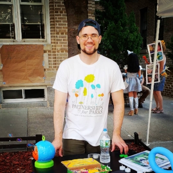 Donnie Welch, young caucasian male stands behind a table in a white T-shirt. On the table are a bubble machine, story book, and supplies for children’s crafts. In the background can be seen apartment buildings and people in costume for  Wizard of Oz  party theme.