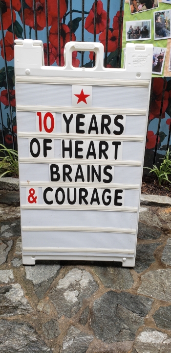 Big white letter board sign with black and red lettering. Sign has a red star at the top and reads “ 10 years / of heart / brains / &amp; courage”. The sign is on a rocky surface with some plants and mulch from the garden visible behind it.