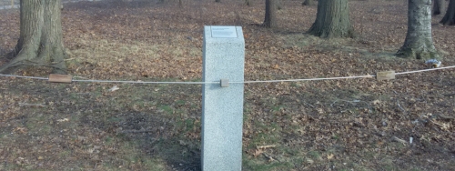 Three wooden objects suspended on a metal string, two rectangles on the left and right and in the middle a cube. A large, cement pole with indistinguishable writing is in the middle of the string behind the cube. Further in the background is a small park with trees and lawn.