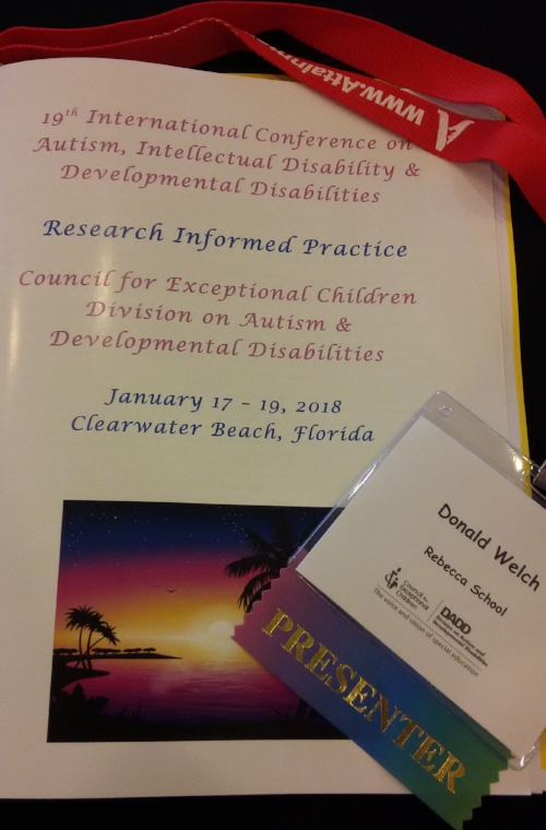 Conference Materials for the CEC-DADD 2018 conference. At top is a red lanyard with a sponsors name on it, the name is blurred by a twist in the lanyard. Below the red lanyard is the program written in blue and pink text on a white, laminated booklet. At the bottom of the booklet is a picture of an island/beach sunset. To the right of this picture is a badge with “Donald Welch” on it and rainbow “presenter” ribbon attached.