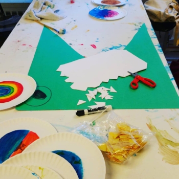 Long white table full of supplies. In the foreground are paper plates with rainbow colors, a bag of yellow cuts and a black expo marker. Next there’s a green poster board cut to look like a crocodile with an eye drawn, on the green poster paper are scissors and white paper cut to look like teeth. The table extends with more rainbow plates in the background.