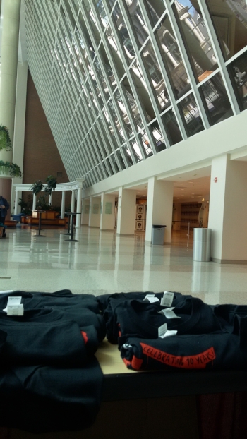 View of the National Housing Center atrium from the merch table. In the foreground, on a table, are rolled up black t-shirts with red stripes bearing black lettering. Beyond that is an atrium with tiled floors and a tall window-like structure jutting out into the atrium space.