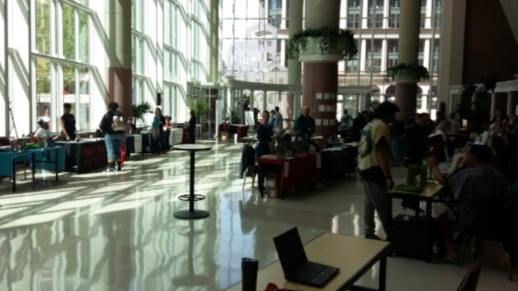 An open room with large windows full of tables, some covered in cloths, other bare, all with materials (books, computers, magazines) on them and with people behind them sitting in chairs or standing up. On the other side, people are walking around and talking to those behind the tables. It’s clear that its sunny outside as light is coming through the large windows of the space and brightening up the book fair scene.