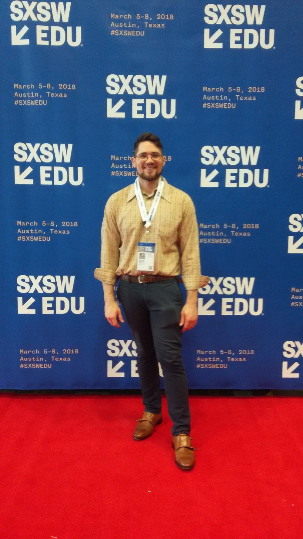 Donnie Welch a young white male with brown hair, a beard, and glasses stands on a red carpet with a blue banner for SXSWedu 2018 behind him. The blue banner his SXSWedu 2018 and the event details in white-yellow hued text. Welch is wearing tan dress shoes, blue pants and a paisley shirt.