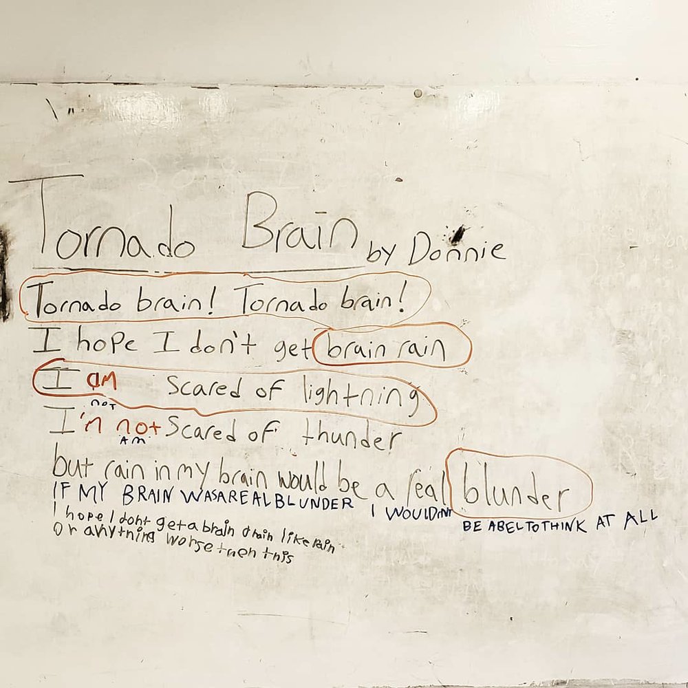 Image of a poem written in black marker on a school white board. Photo editing has given everything a slight burnt tint. The poem’s title says “Tornado Brain” by Donnie. Parts of the poem are circled in orange, and brown other lines have writing around them in blue and purple ink.