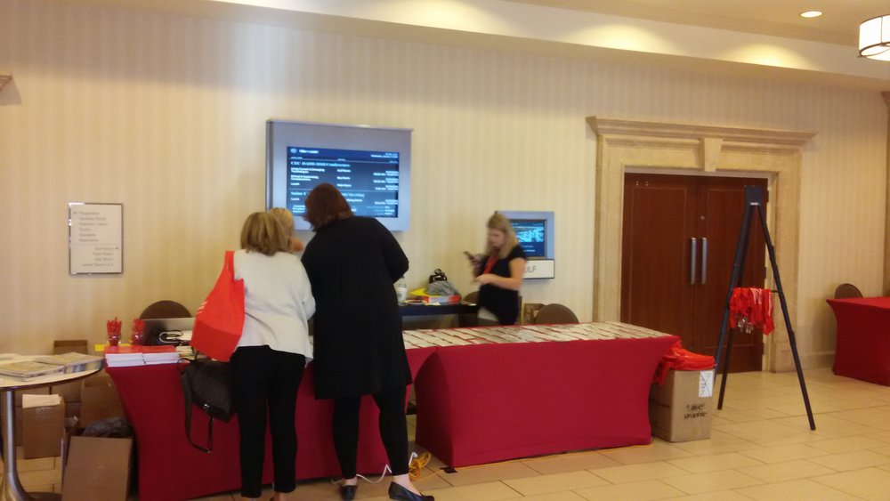 Table at the CEC-DADD 2018 conference. Two older Caucasian women look at pamphlets on a red clothed table. A Third middle-aged Caucasian women is seen checking her phone behind the table.
