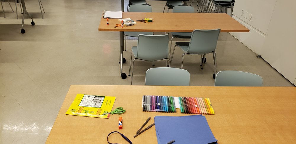 Two wooden tables with books and supplies on them. The front table has colored felt pens, blue construction paper, scissors, glue sticks, pens, and a yellow children’s book. The back table has lined paper, crayons, scissors, glue, and a full length book. Both tables have gray chairs around them.