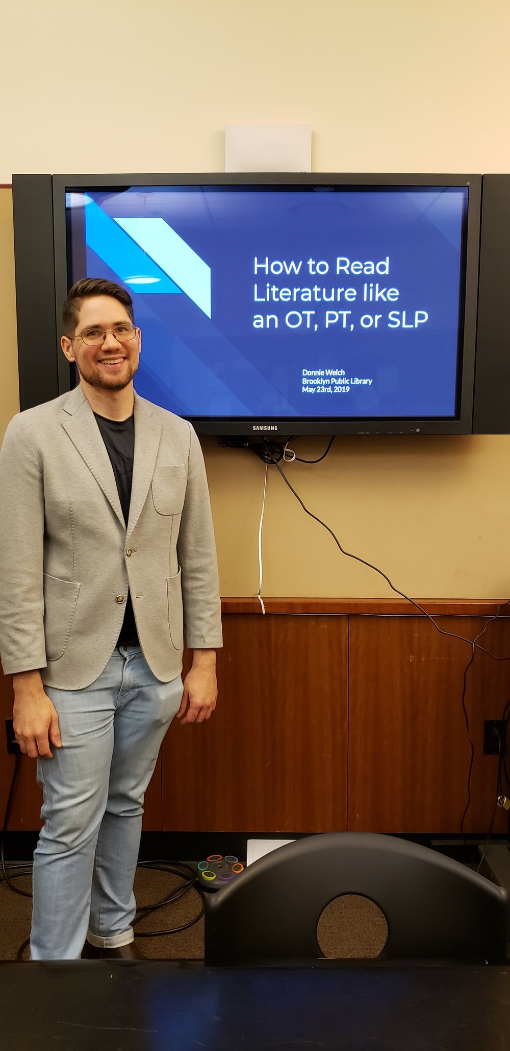 Donnie Welch, a young white man, stands smiling at the front of a room in business casual attire. Behind him on a large, computer monitor is a presentation title slide reading, “How to Read Literature like an OT, PT, or SLP” in white lettering on a blue background.