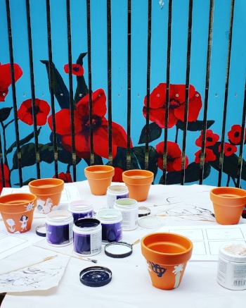 Orange/Brown ceramic pots on a white table cloth with various shades of paint in purple and white pots. Some paintings are visible on the pots, others appear blank. In the background is a vibrant teal slatted fence with red poppies painted on.