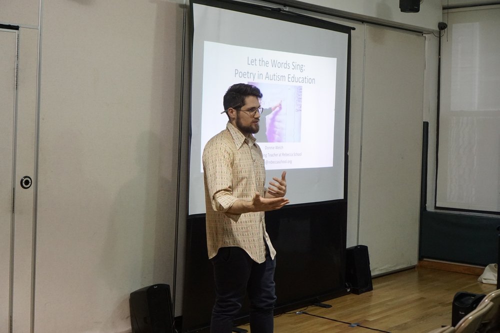 Donnie Welch, a young bearded man with glasses, speaks in a school auditorium, gesturing with his hands out. Next to him is a projector displaying a slide on a screen him.