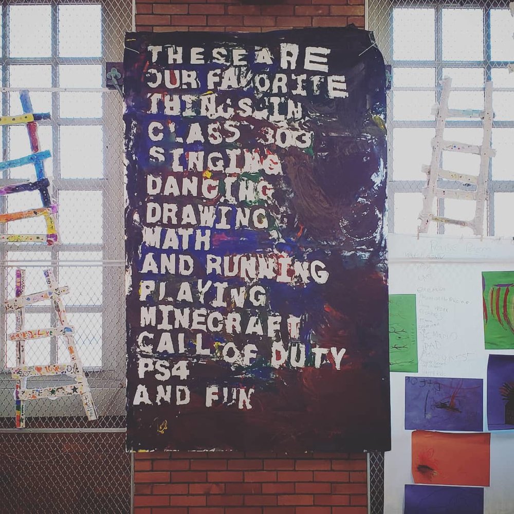 The poem hanging up on a brick school wall. Other pieces of art from other projects surround it: individual drawings and crafted ladders. The paint colors (red, blue, brown, black, yellow, green) have dried. The poem reads “These are / our favorite / things in / class 303 / Singing / Dancing / Drawing / Math / And running / playing Minecraft / call of duty / PS4 / and Fun”