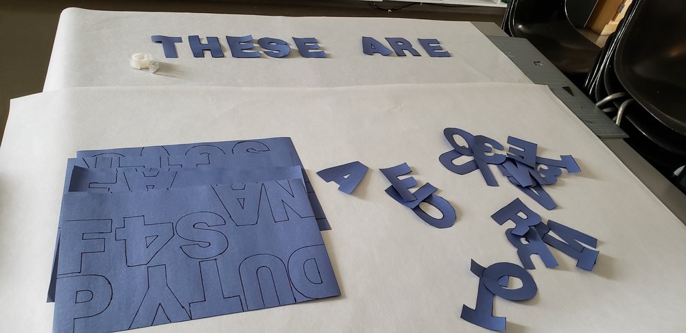 Large white mural paper. “These Are'“ spelled out at the top in blue letters cut from the construction paper. Below there are more pieces of paper with tracings and a pile of letters cut out not yet organized into words.