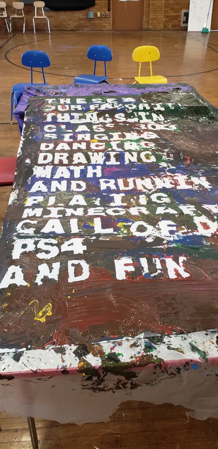 Poem laid out on the tables, but now the white mural paper is covered in various layers of paints: black, green, blue, pink, brown, red, and yellow. The blue construction paper letters have been removed so that now the letters come through from the empty space, white gaps on the mural paper. The poem reads The poem reads “These are / our favorite / things in / class 303 / Singing / Dancing / Drawing / Math / And running / playing Minecraft / call of duty / PS4 / and Fun”