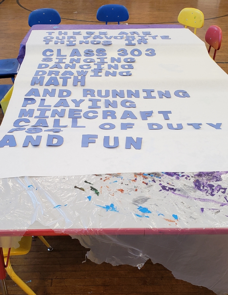 White mural paper with the poem in blue construction paper letters laid out on tables covered in plastic. The poem reads “These are / our favorite / things in / class 303 / Singing / Dancing / Drawing / Math / And running / playing Minecraft / call of duty / PS4 / and Fun”