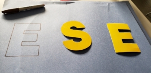 Three letter “E” “S” “E”on blue construction paper. One of the letters is an outline in sharpie, the other two are yellow stencils. The sharpie is seen in the top left corner of the paper.