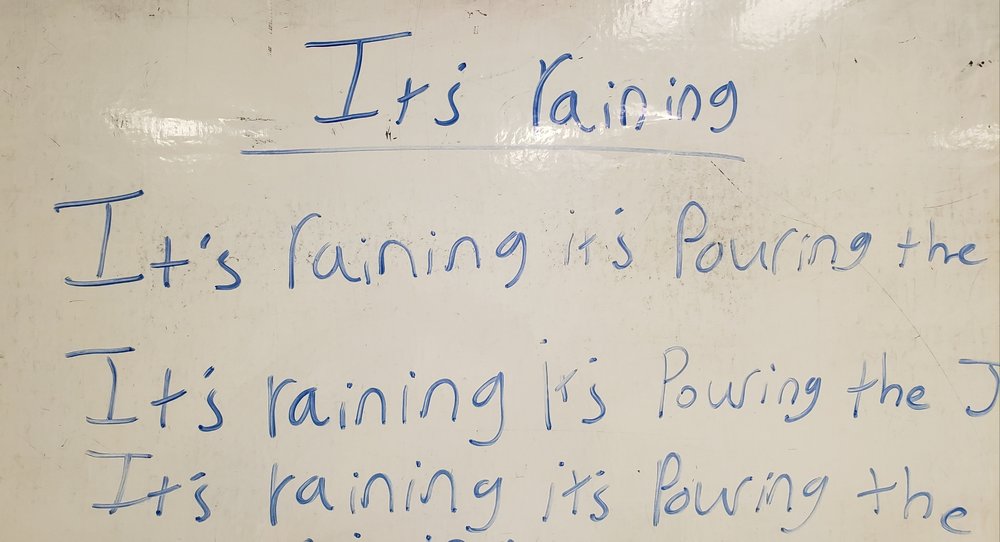 Words on a white board in blue expo marker read “It’s raining it’s pouring the” This line is repeated again and again.