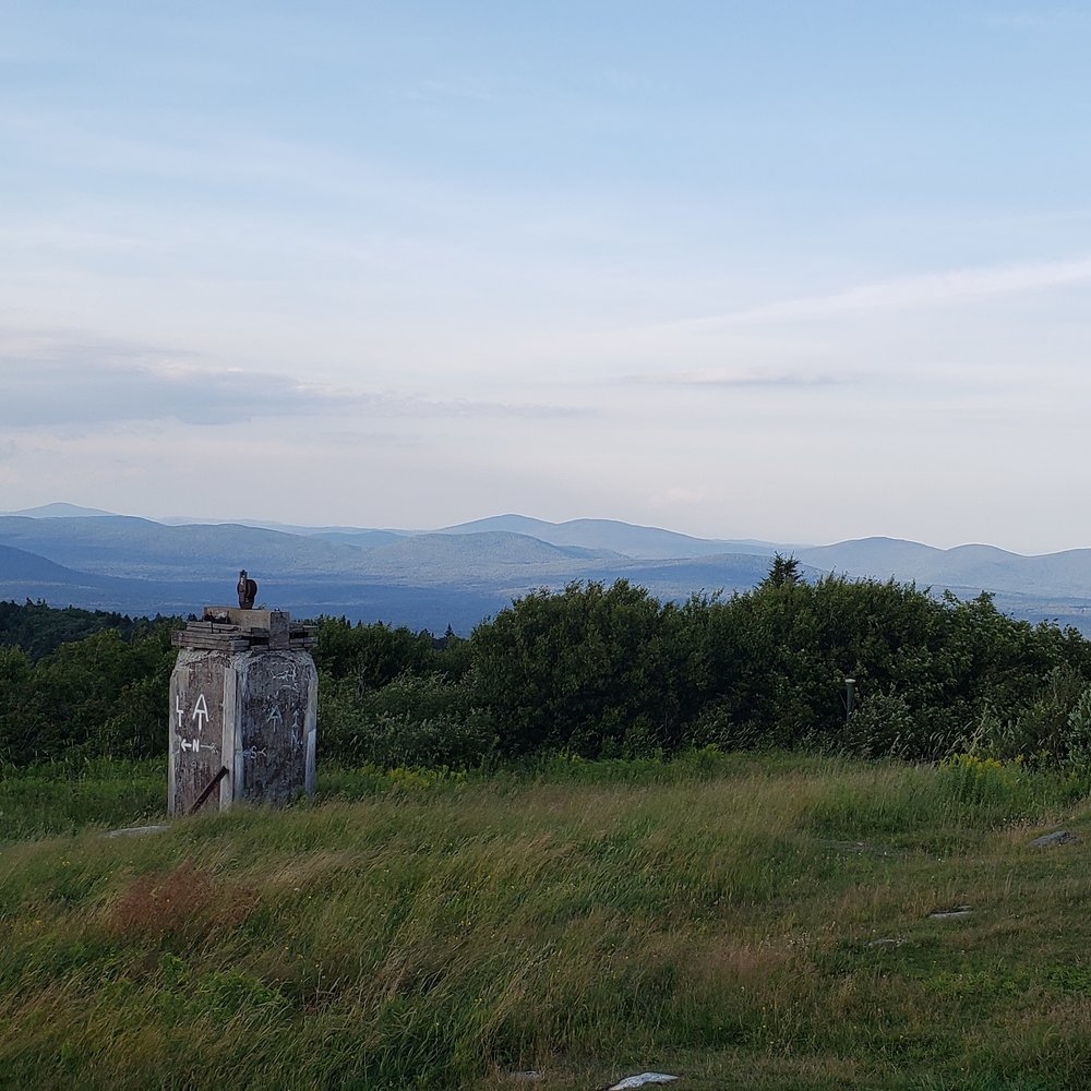 Sunset view off Bromley Mountain. In the foreground is a field of tall grass bent by the wind and an old stone work with LT and AT symbols and white N for North painted on in white.In the background can be seen ranges of mountains pale blue and purple from the light at sunset.