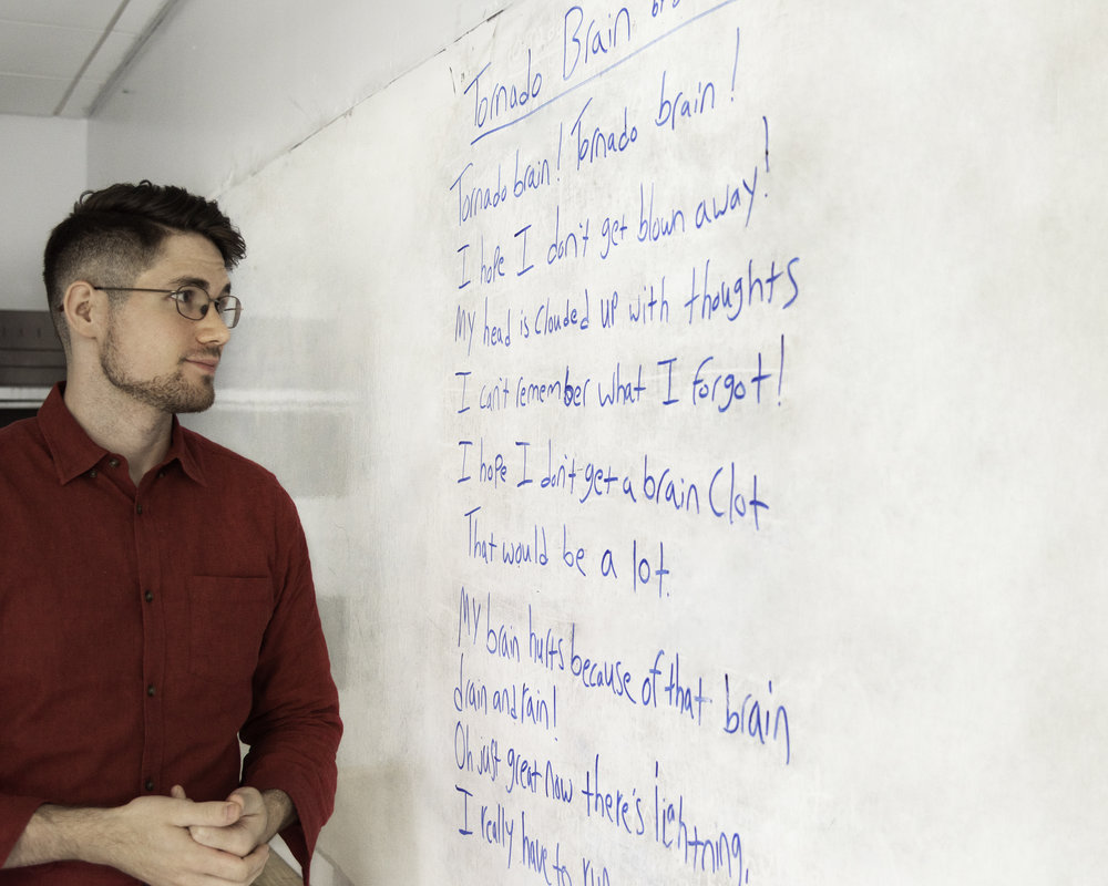 Donnie Welch, a young white man with glasses wearing a red button-down shirt, looks at a classroom white board with his hands clasped. On the board is some writing from an inclusive poetry workshop in blue expo. The angle of the photo makes it difficult to read some of the text, but it’s structured in a poetic manner with line breaks.