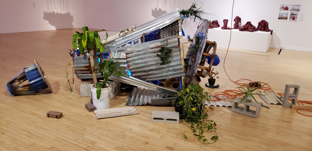 Contemporary art installation on a hardwood floor. The art work looks like a 1970’s space capsule, but covered in sheet metal, plants, and bricks. Extension cables can be seen running out of the piece as well. In the background are other photos and statues.