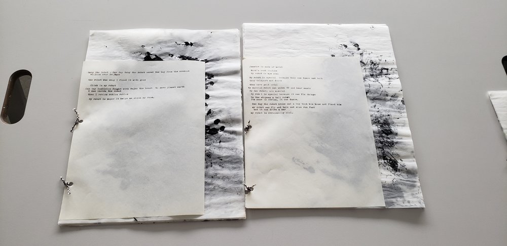 Two books handmade books side-by-side on a table. Both are tied together by a bit of twine, the front of them have typewritten words that are indistinguishable at the photo’s distance. Behind these front pages are pieces of tracing paper with blotches and dots of black paint.