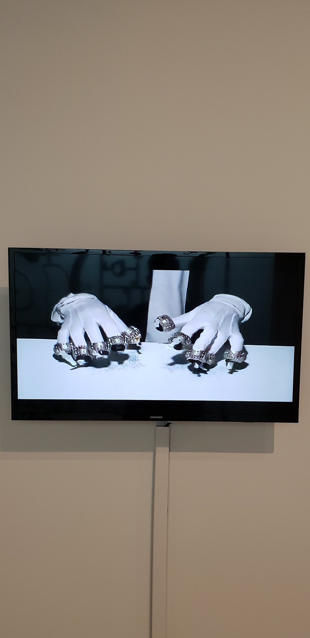 On a flatscreen TV gloved hands can be seen with metallic tips on the ends of each fingers. The hands seem to be typing with small, black residue visible underneath the fingers.