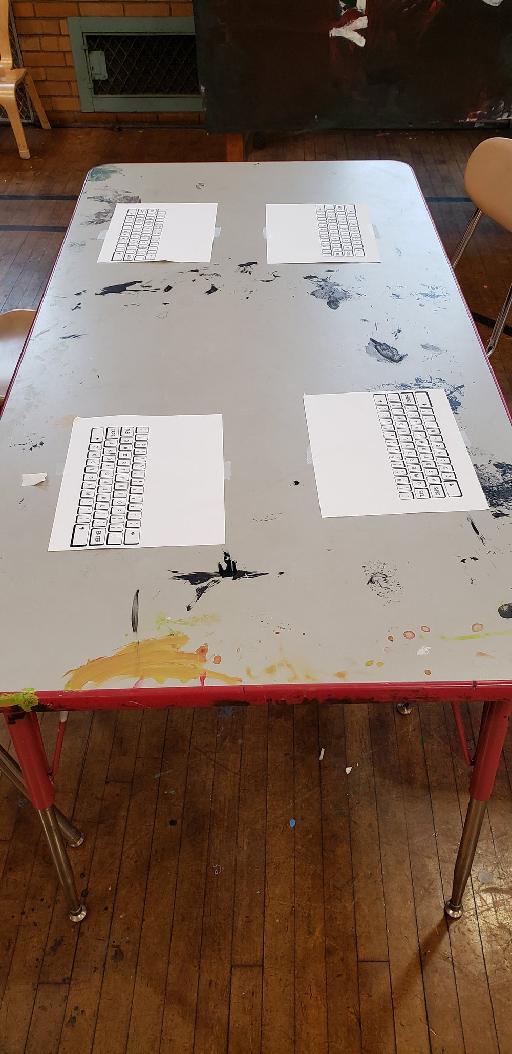 On an art room table, covered in old paint marks, print outs of computer keyboards are taped down.