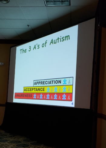 Large presentation screen with a black curtain skirt on the bottom. On the top in Green is “ The 3 A’s of Autism” below is a stack of three words showing each concept as a foundation for the other. From the bottom up in red “Awareness” in yellow “Acceptance” and in light blue “Appreciation”: