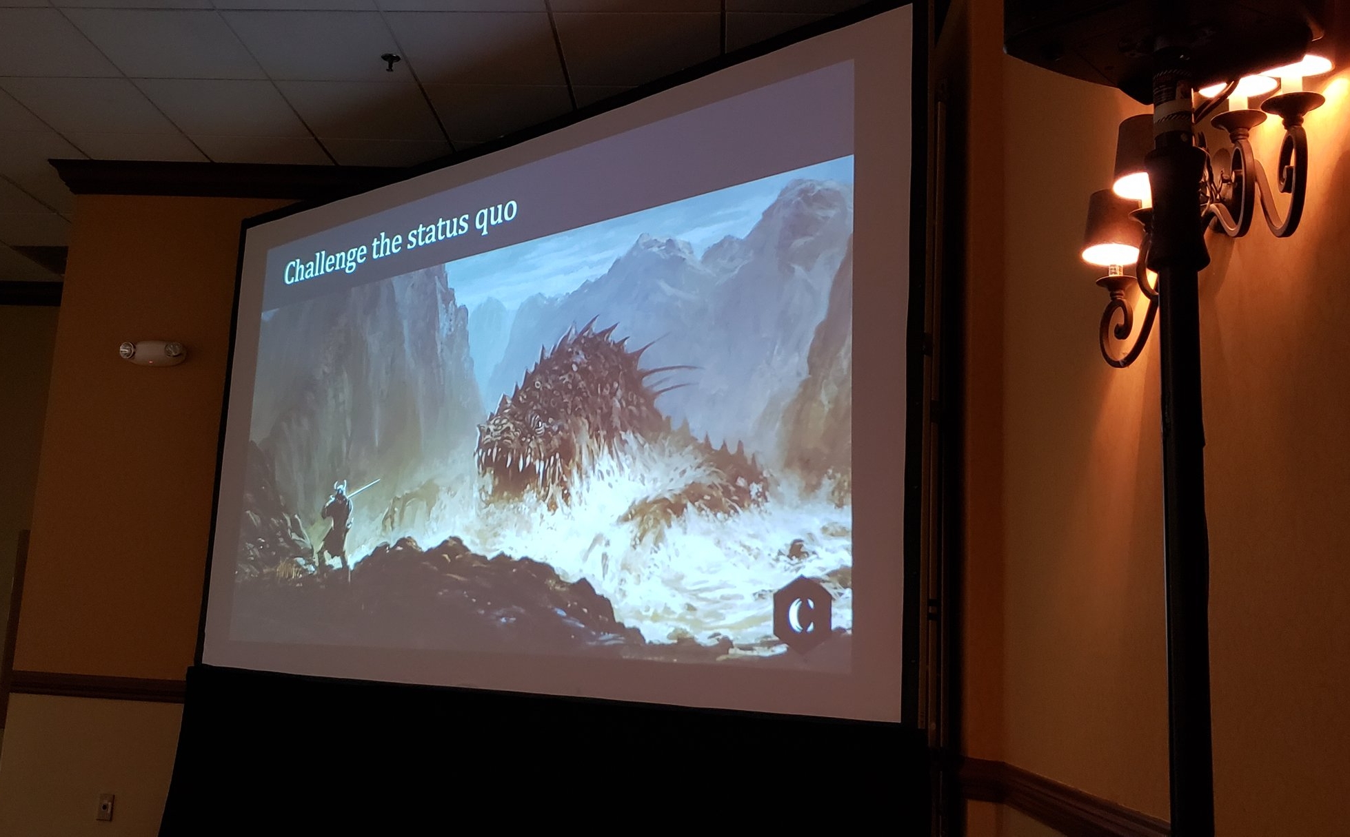 Large presentation screen with black, curtain skirt. On top of the screen in large white lettering “Challenge the Status Quo” Under that is an image of a fantasy, warrior character facing a large dragon-like monster emerging from a body of water. In the bottom right is the C logo for Critical Core.
