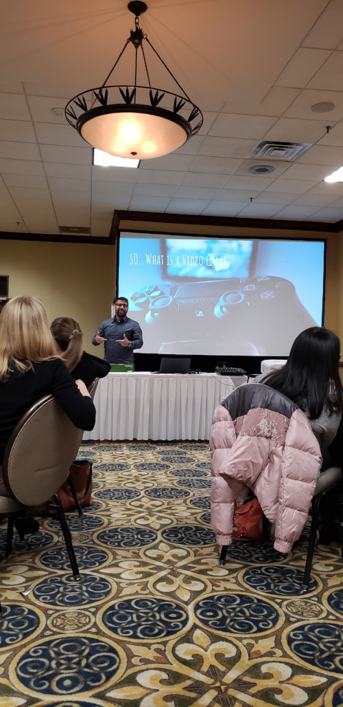 Chris Hernandez a young Puerto-Rican man, presents from behind a table to a group of people sitting. His body language suggests he is engaged and mid-speech. Behind him is a large presentation screen with “So…What is a Video Game” written over the image of a console controller. Around Hernandez can be seen equipment such as a laptop, sound board, and projector. The presentation is taking place in the hotel ballroom and the navy and yellow rug and yellow wall of the room are seen in the photo.