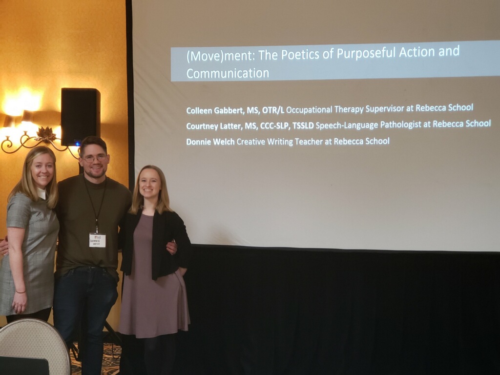 From left to right: Colleen Gabbert, young white woman. Donnie Welch young white man. Courtney Latter young white woman. All dressed professionally. The three are posing in front of their title slide on a large presentation screen with a black, curtain skirt. Behind them can be seen a speaker and light as well as the navy-yellow rug and yellow wall of the hotel ballroom.