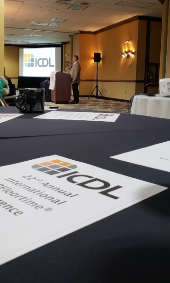 Jeff Guenzel a middle aged white male with professionally dressed addresses an audience from behind a podium. In the foreground is a table with a conference flyer and in the background, behind Guenzel, is the ICDL logo on a power point screen. Around Guenzel can be seen equipment such as projector, speakers, lights. The walls of the hotel lobby are yellow and the floor has a navy and yellow rug to match.
