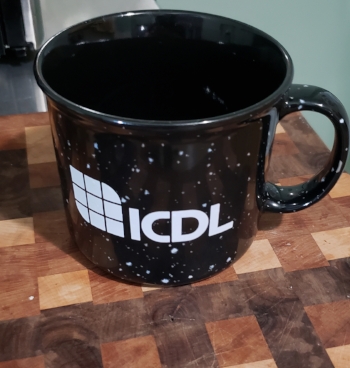 Black and white ceramic style coffee mug with “ICDL" and ICDL’s Logo in bold white lettering on the front. The coffee mug is sitting on a checkered wooden cutting board.