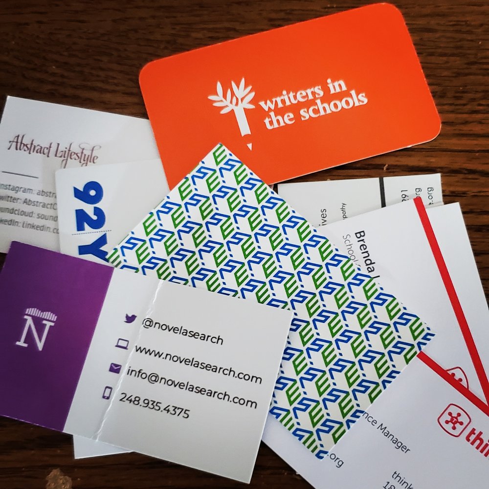 Pile of business cards, mostly white with colored text and images, on top of a brown wooden desk.