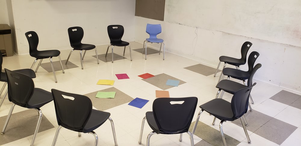 A circle of classrrom chairs, black and one blue, on a tiled floor. In the middle of the circle of chairs is a smaller circle of colored cards. In the foreground is a classroom dry-erase whiteboard.