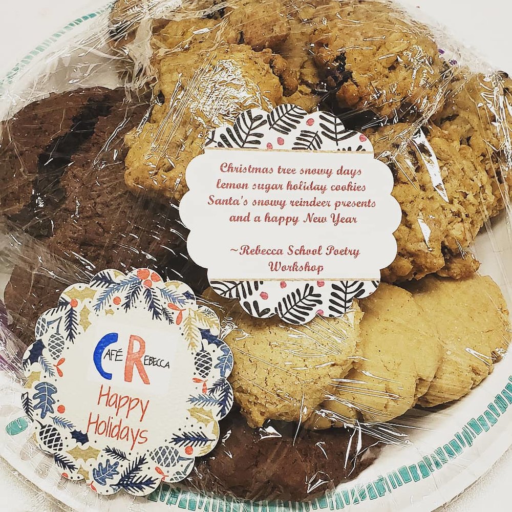 Pile of three flavored cookies, chocolate, sugar, and oatmeal on a plastic wrapped plate. On top of the plate are two festive tags. One tag shows the front with the Cafe Rebecca logo, the other tag shows the back with the Rebecca School Poetry Workshop’s poem.