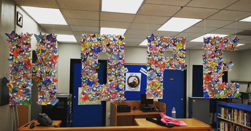 The word "hope" spelled out in large, capital letters cut from poster paper and suspended in a school library. Inside the letters are butterflies of various colors crafted out of paper.