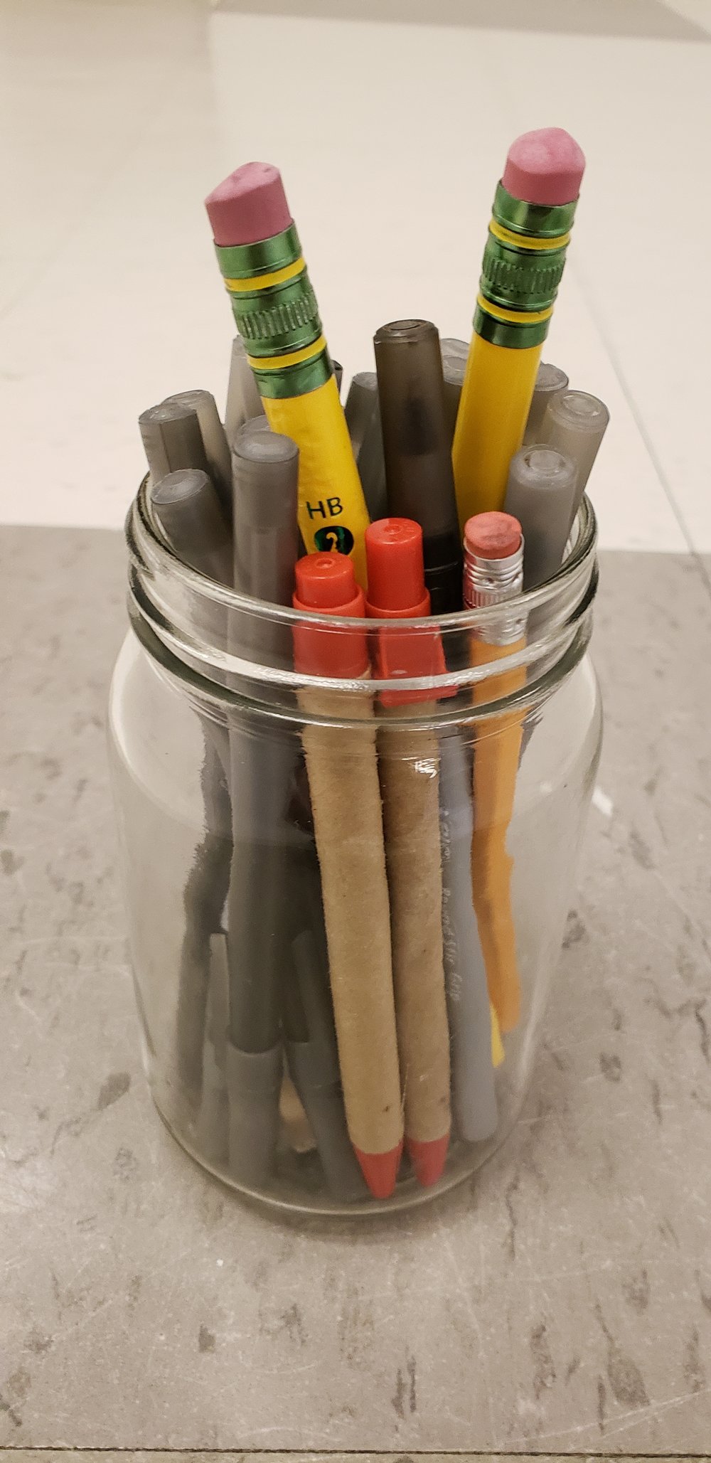 Clear mason jar of writing utensils on a tiled classroom floor. The Jar has plastic ball point pens with both smooth and rough grips, pens with cardboard wrapping, regular sized pencils, and giant pencils.