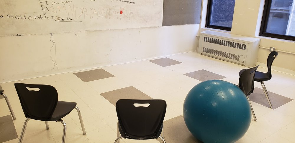 Tiled classroom with a white board. Black chairs arranged in a half-circle around the white board. In the middle of the chairs is a teal yoga ball intended to be a seat. On the board can be seen some writing.